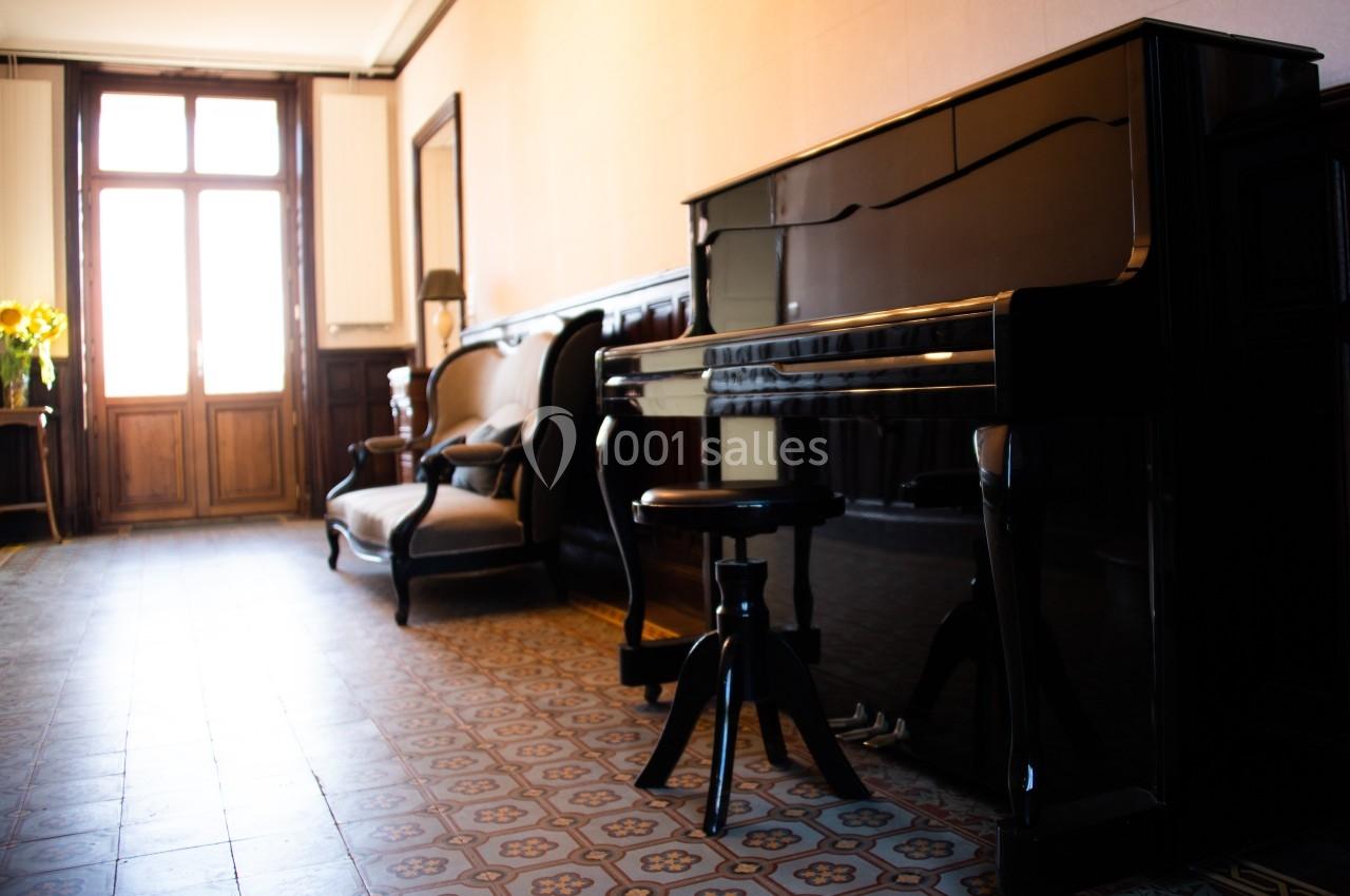 Piano droit noir dans un salon lumineux avec carrelage ancien, fauteuils en bois et une fenêtre en arrière-plan.