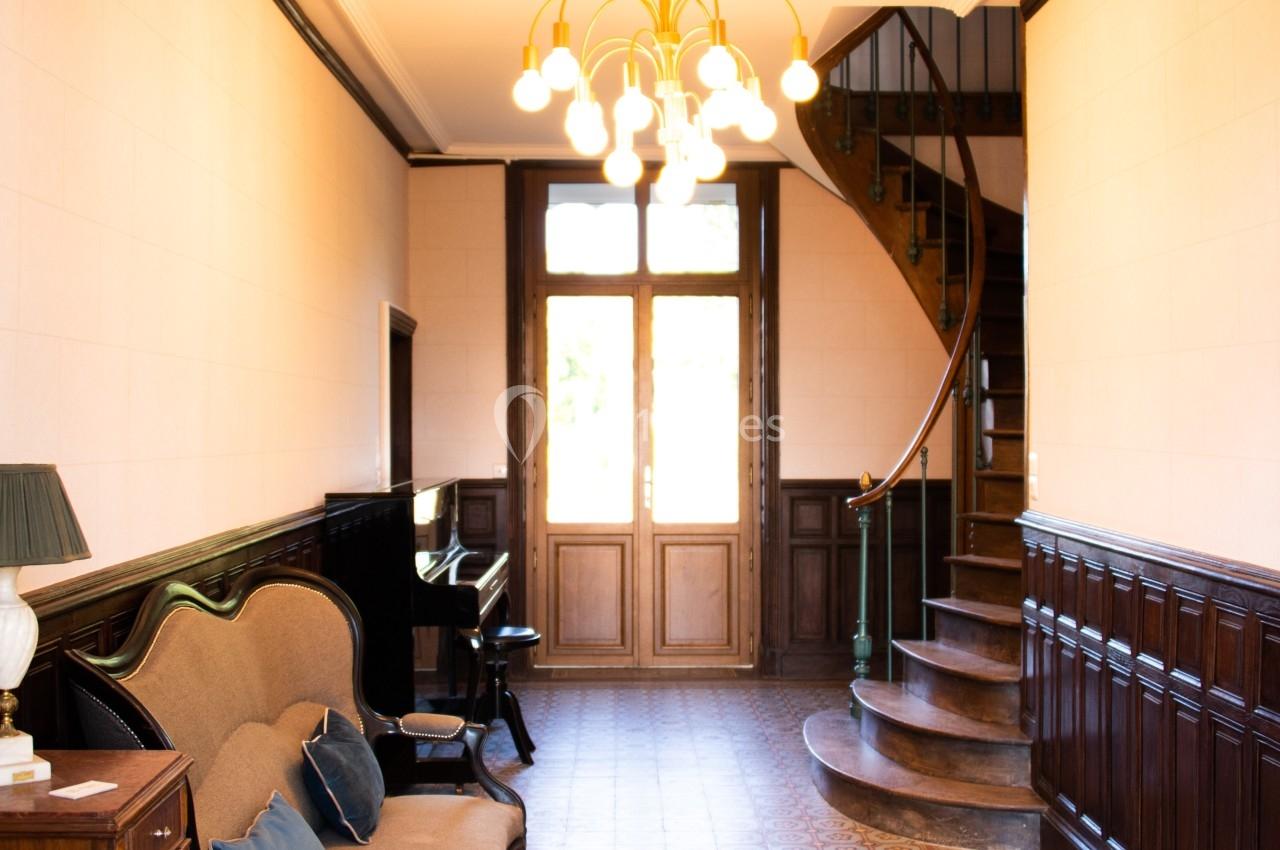 Couloir lumineux avec carrelage ancien, canapé, piano, escalier en colimaçon et porte-fenêtre en arrière-plan.