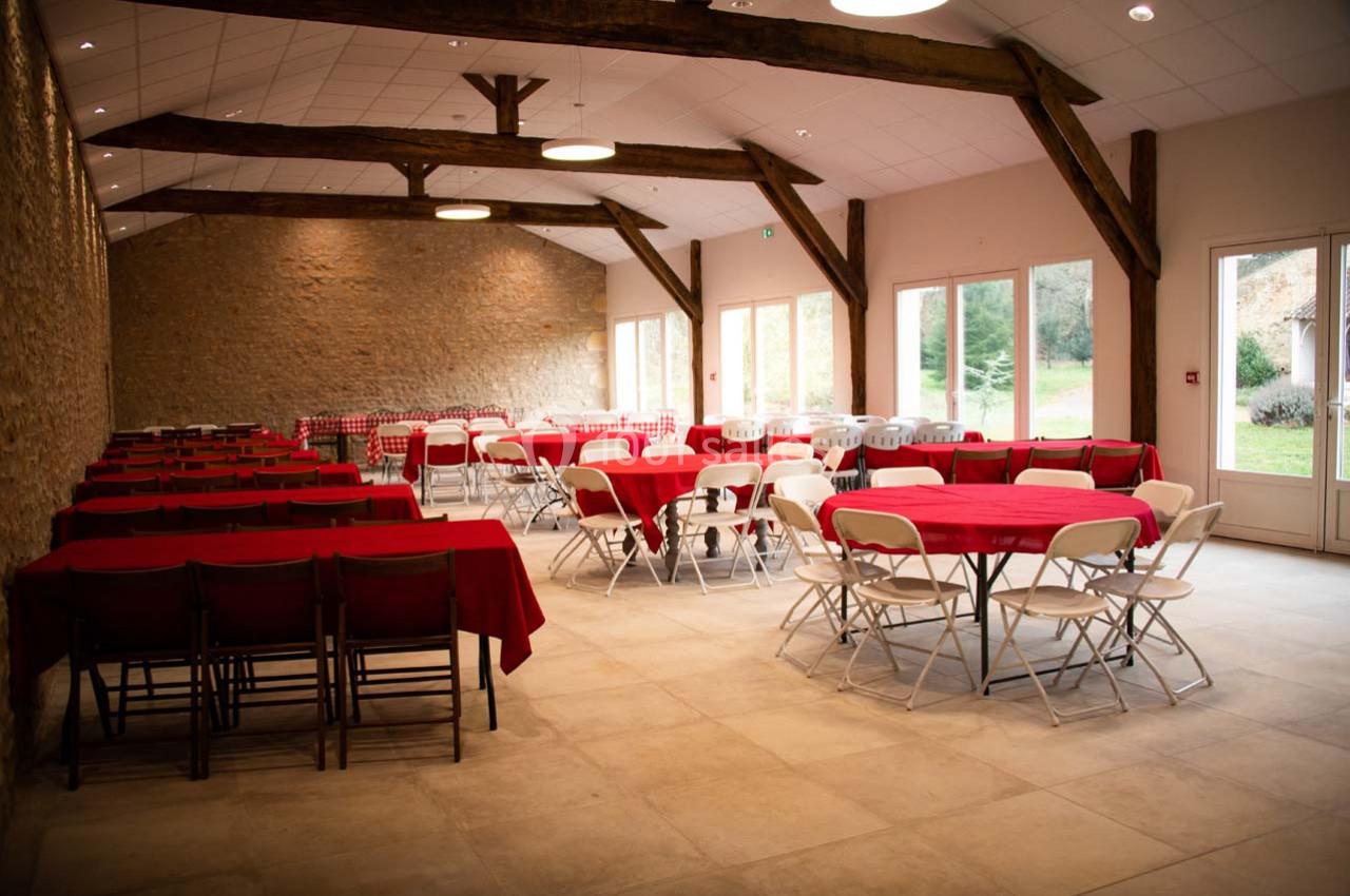 Salle lumineuse avec murs en pierre, tables recouvertes de nappes rouges et chaises blanches, vue sur un jardin.