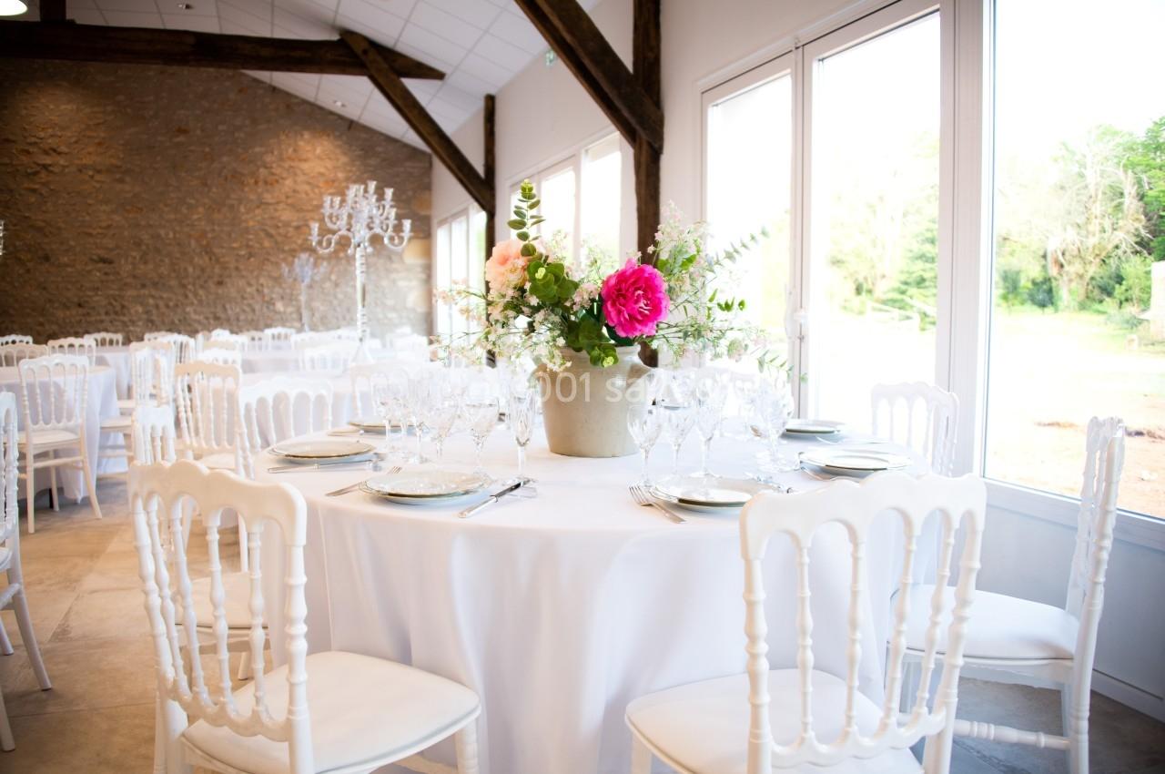 Salle de réception lumineuse avec tables rondes dressées, décorées de fleurs dans un vase central.