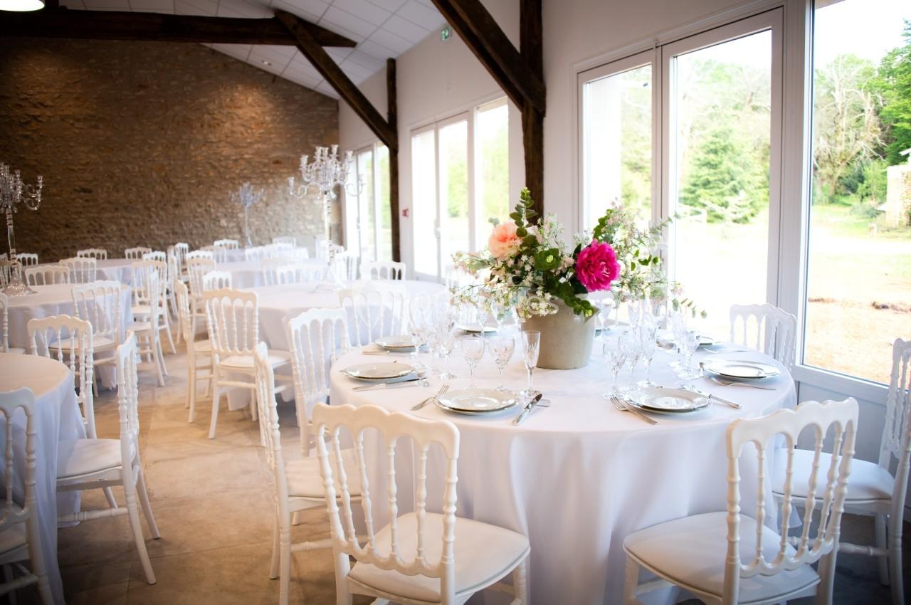 Salle de réception lumineuse avec tables rondes dressées, chaises blanches et centre de table floral.