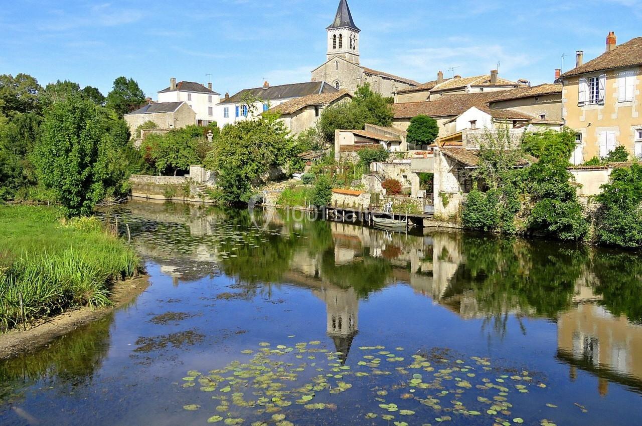 Vue d'un village avec une église, des maisons en pierre et une rivière bordée de végétation, reflet dans l'eau calme.