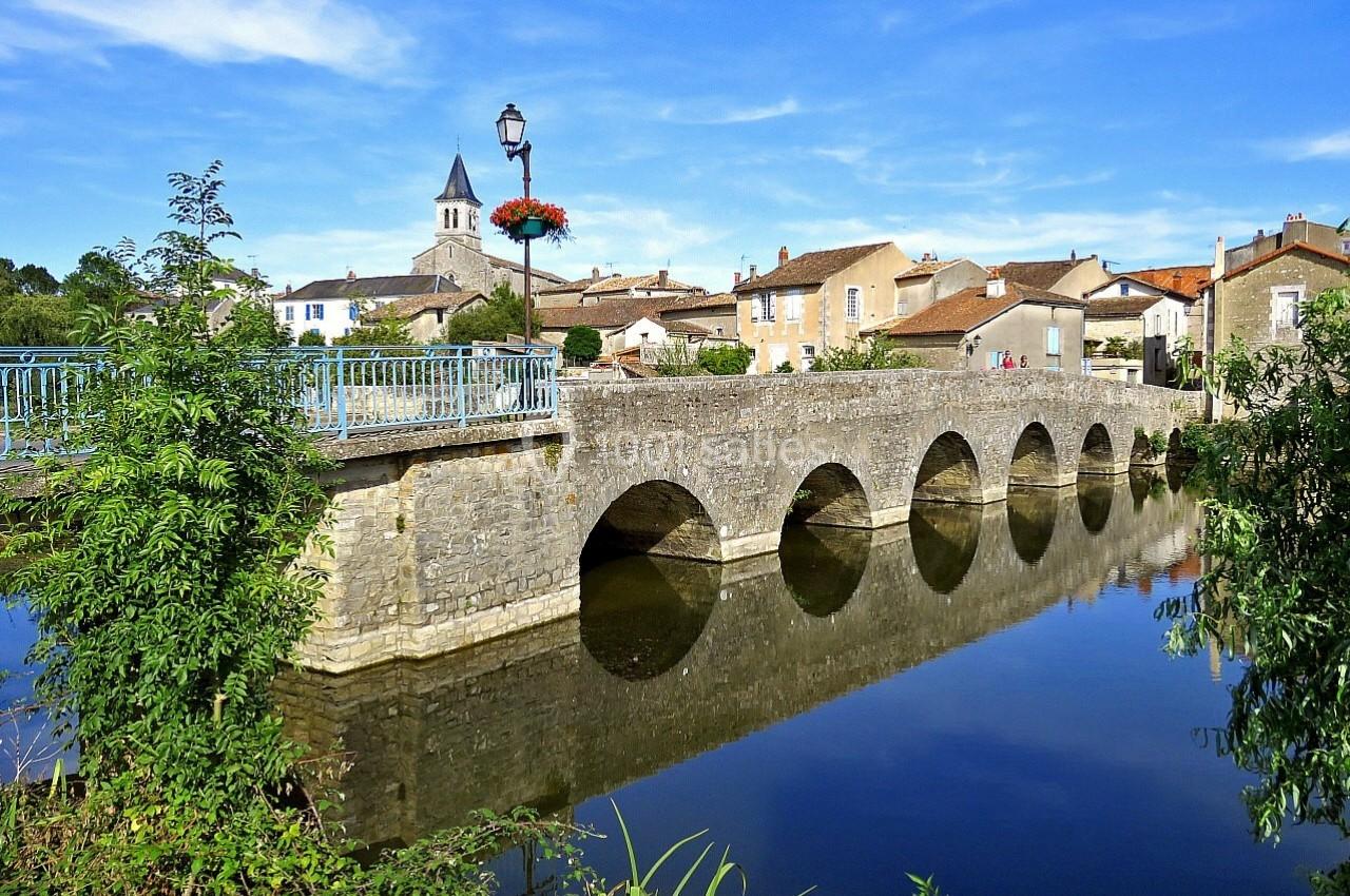 Vue d'un pont en pierre à arches traversant une rivière, avec un village et un clocher en arrière-plan.