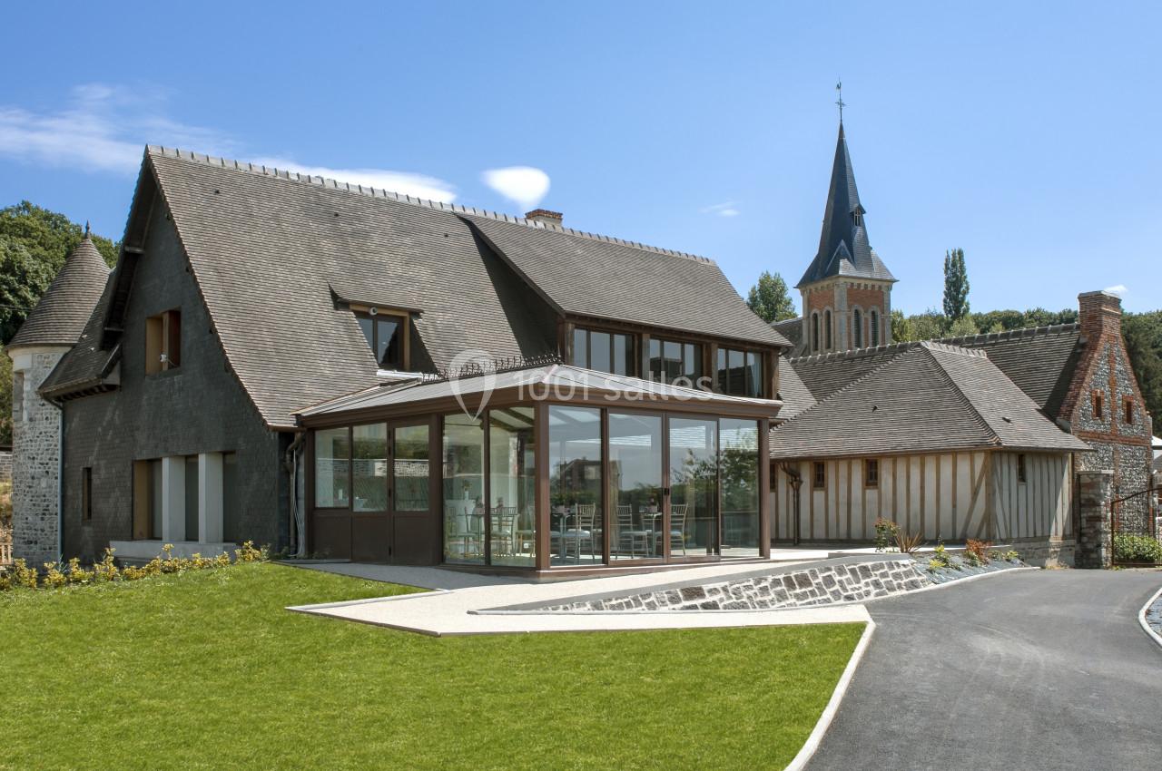 Location salle Saint-Samson-de-la-Roque (Eure) - Manoir Néo Normand #2 Grande maison en pierre avec extension vitrée, jardin aménagé et clocher d'église visible en arrière-plan.