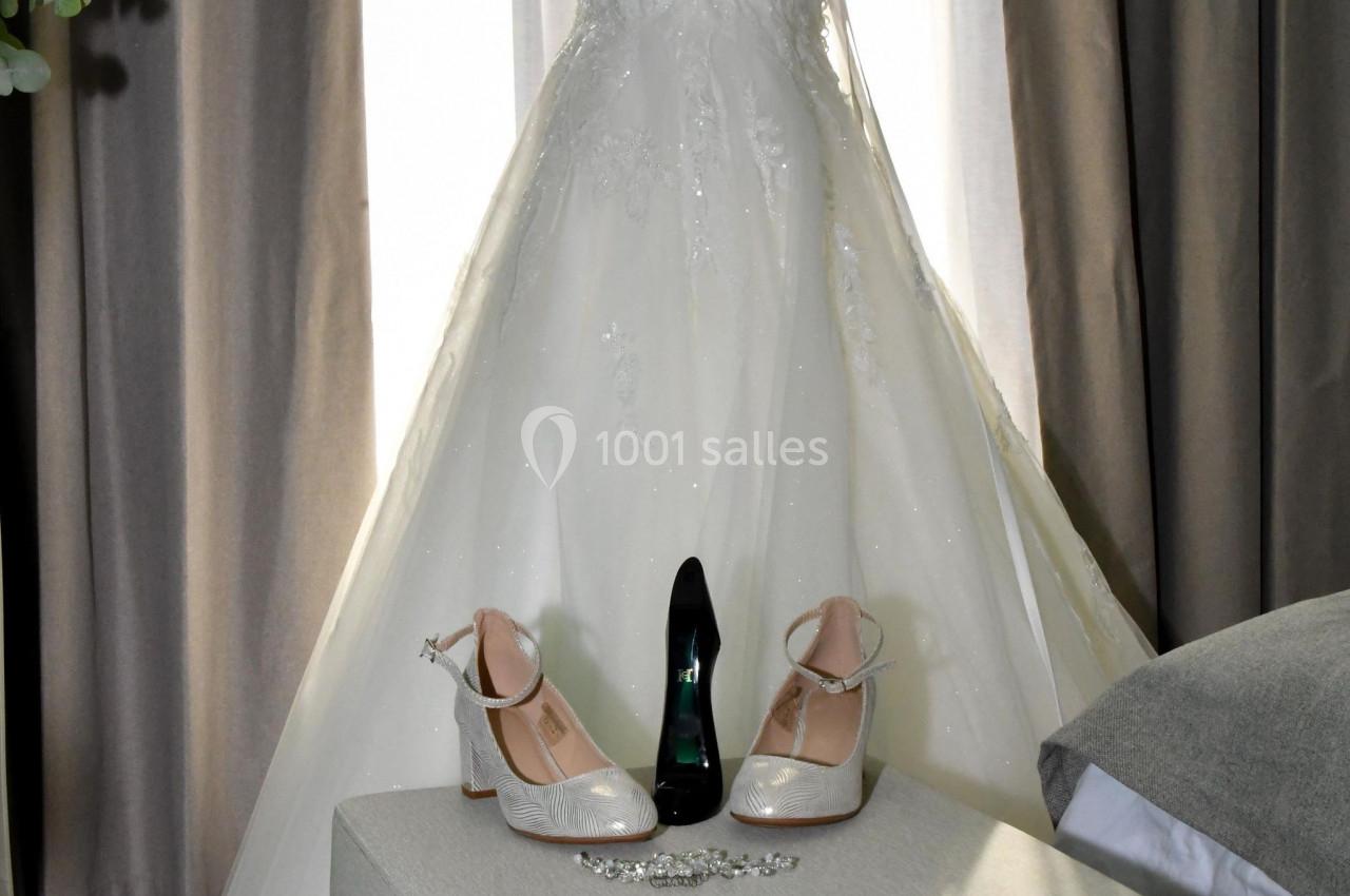 Location salle Saint-Samson-de-la-Roque (Eure) - Manoir Néo Normand #64 Robe de mariée blanche sur un mannequin, entourée de chaussures, d'un bijou et d'un rideau en arrière-plan.