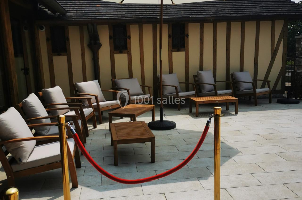 Location salle Saint-Samson-de-la-Roque (Eure) - Manoir Néo Normand #21 Terrasse ensoleillée avec mobilier en bois et coussins, parasol central et cordons rouges délimitant l'espace.