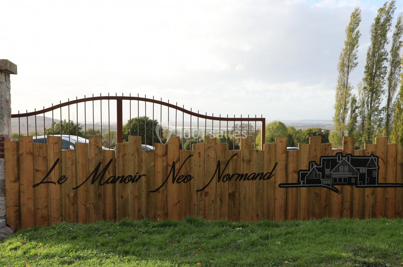 Location salle Saint-Samson-de-la-Roque (Eure) - Manoir Néo Normand #30 Clôture en bois avec le texte ’Le Manoir Néo Normand’ et un dessin de maison, devant un paysage rural.