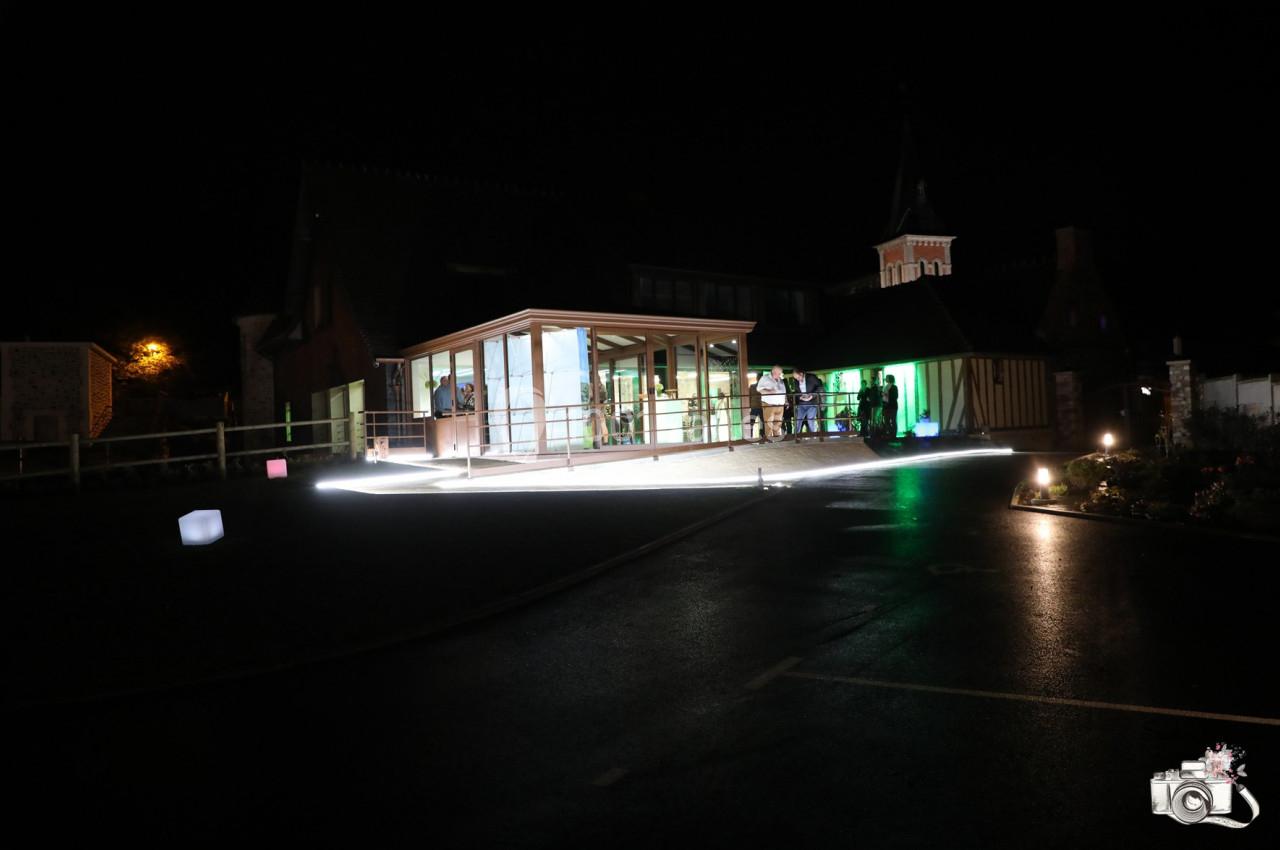 Location salle Saint-Samson-de-la-Roque (Eure) - Manoir Néo Normand #54 Bâtiment éclairé la nuit avec des lumières colorées, entouré d'une cour sombre et de quelques personnes visibles à l…