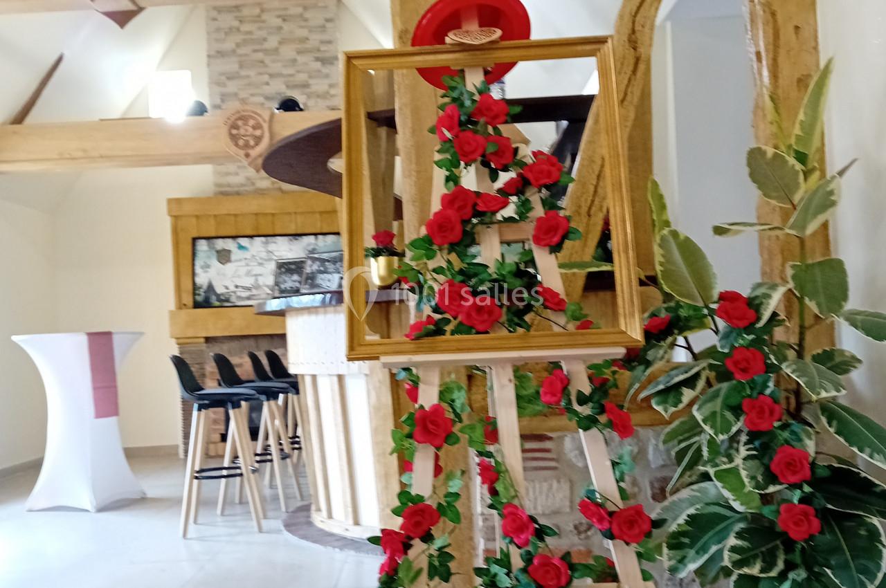 Location salle Saint-Samson-de-la-Roque (Eure) - Manoir Néo Normand #56 Chevalet décoré de roses rouges dans une salle avec poutres en bois, bar et plantes en arrière-plan.