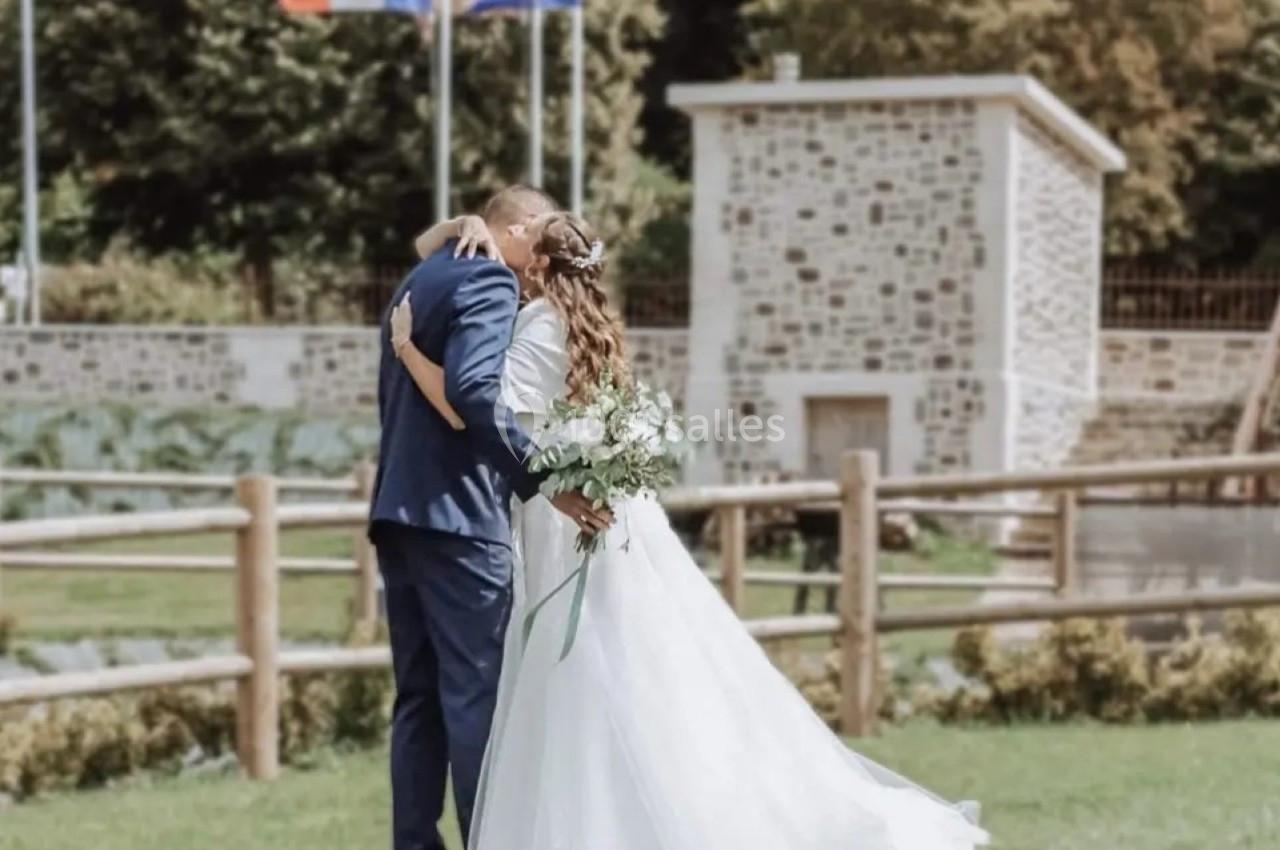 Location salle Saint-Samson-de-la-Roque (Eure) - Manoir Néo Normand #9 Un couple en tenue de mariage s'embrasse dans un jardin, avec des drapeaux et un bâtiment en pierre en arrière-plan.