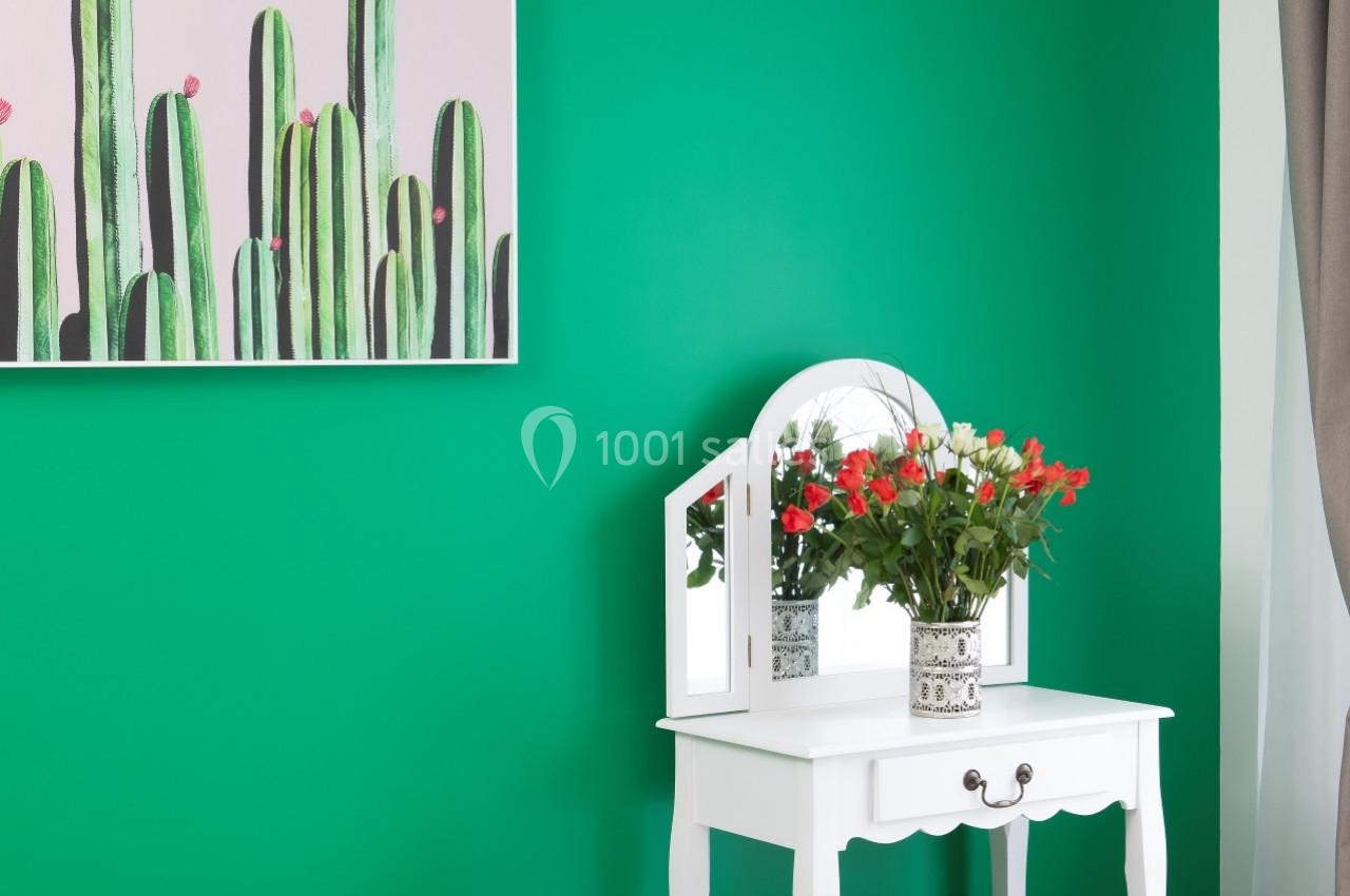 Location salle Saint-Samson-de-la-Roque (Eure) - Manoir Néo Normand #65 Coiffeuse blanche avec miroir, vase de fleurs et tabouret, devant un mur vert décoré d'un tableau de cactus.