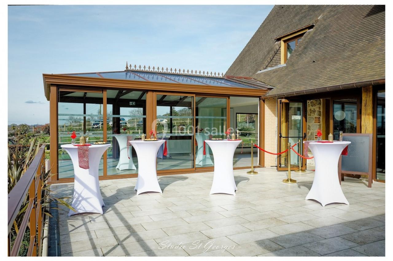 Location salle Saint-Samson-de-la-Roque (Eure) - Manoir Néo Normand #72 Terrasse avec tables hautes drapées de blanc, décorées de fleurs rouges, devant une salle vitrée.