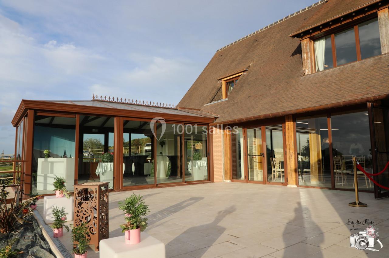 Location salle Saint-Samson-de-la-Roque (Eure) - Manoir Néo Normand #90 Terrasse ensoleillée avec vue sur une salle vitrée, entourée de plantes et d'une architecture en bois et briques.