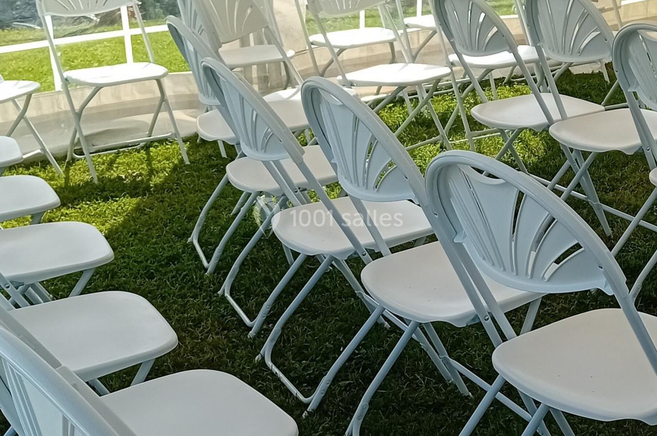 Location salle Saint-Samson-de-la-Roque (Eure) - Manoir Néo Normand #101 Chaises blanches pliantes alignées sous une tente, posées sur une pelouse verte par une journée ensoleillée.