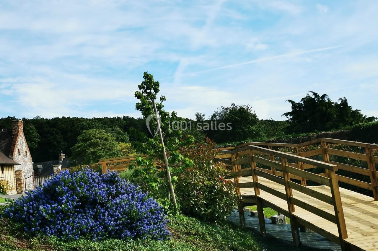 Location salle Saint-Samson-de-la-Roque (Eure) - Manoir Néo Normand #103 Passerelle en bois traversant un jardin verdoyant avec des fleurs bleues et une maison en arrière-plan.