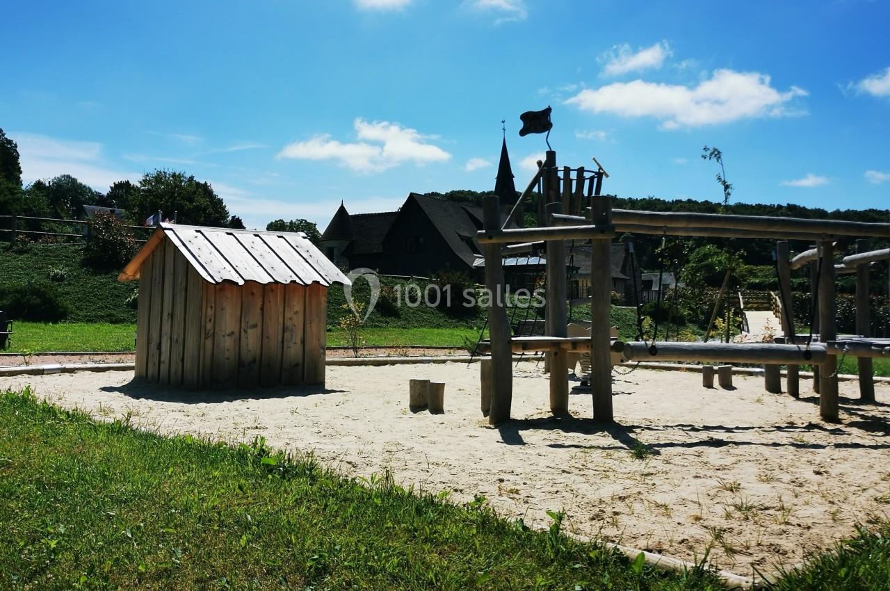 Location salle Saint-Samson-de-la-Roque (Eure) - Manoir Néo Normand #105 Aire de jeux en bois sur sol sablé, entourée de verdure et de bâtiments en arrière-plan sous un ciel bleu.