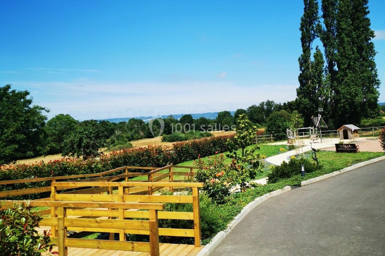 Location salle Saint-Samson-de-la-Roque (Eure) - Manoir Néo Normand #106 Passerelle en bois surplombant un jardin fleuri avec des arbres et un ciel dégagé en arrière-plan.
