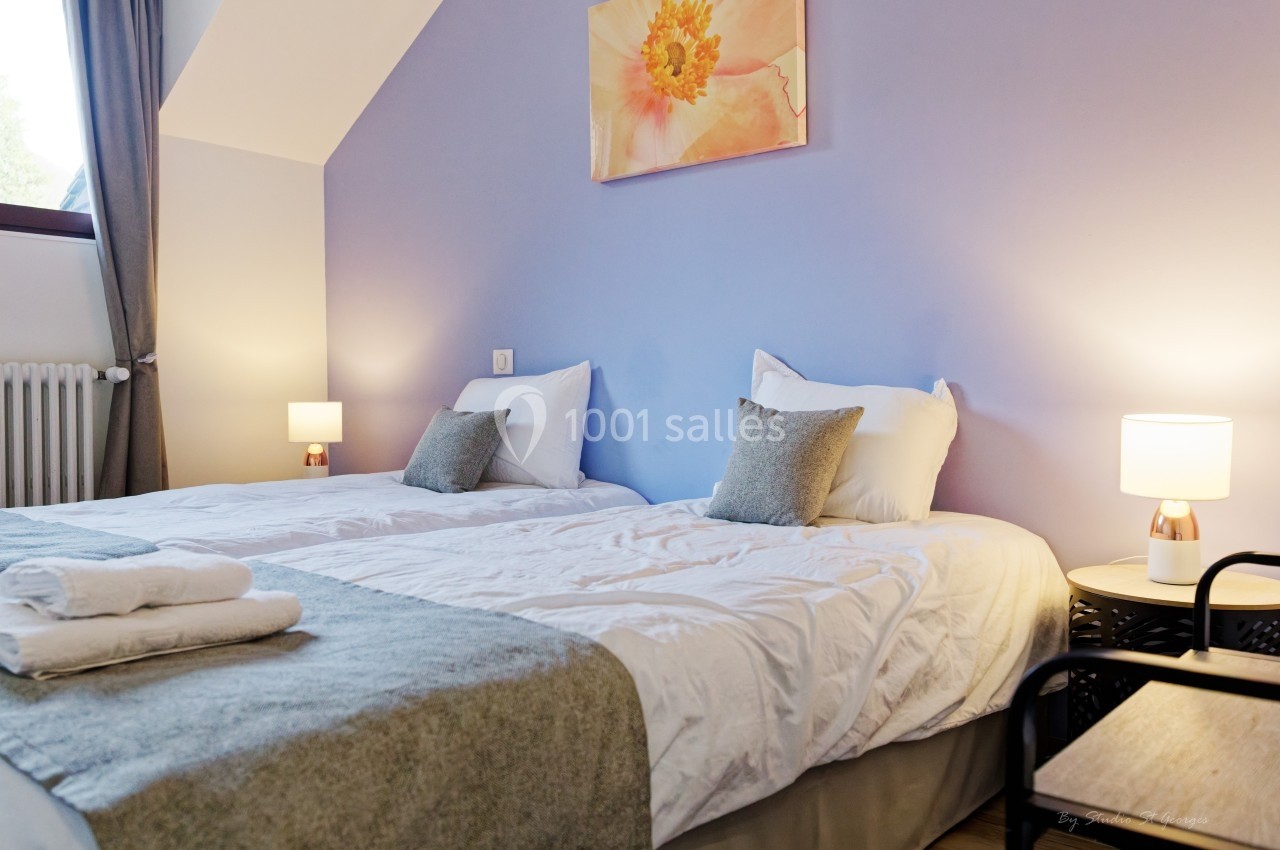 Deux lits simples avec linge de lit blanc, table de chevet et lampe, dans une chambre aux murs violets.