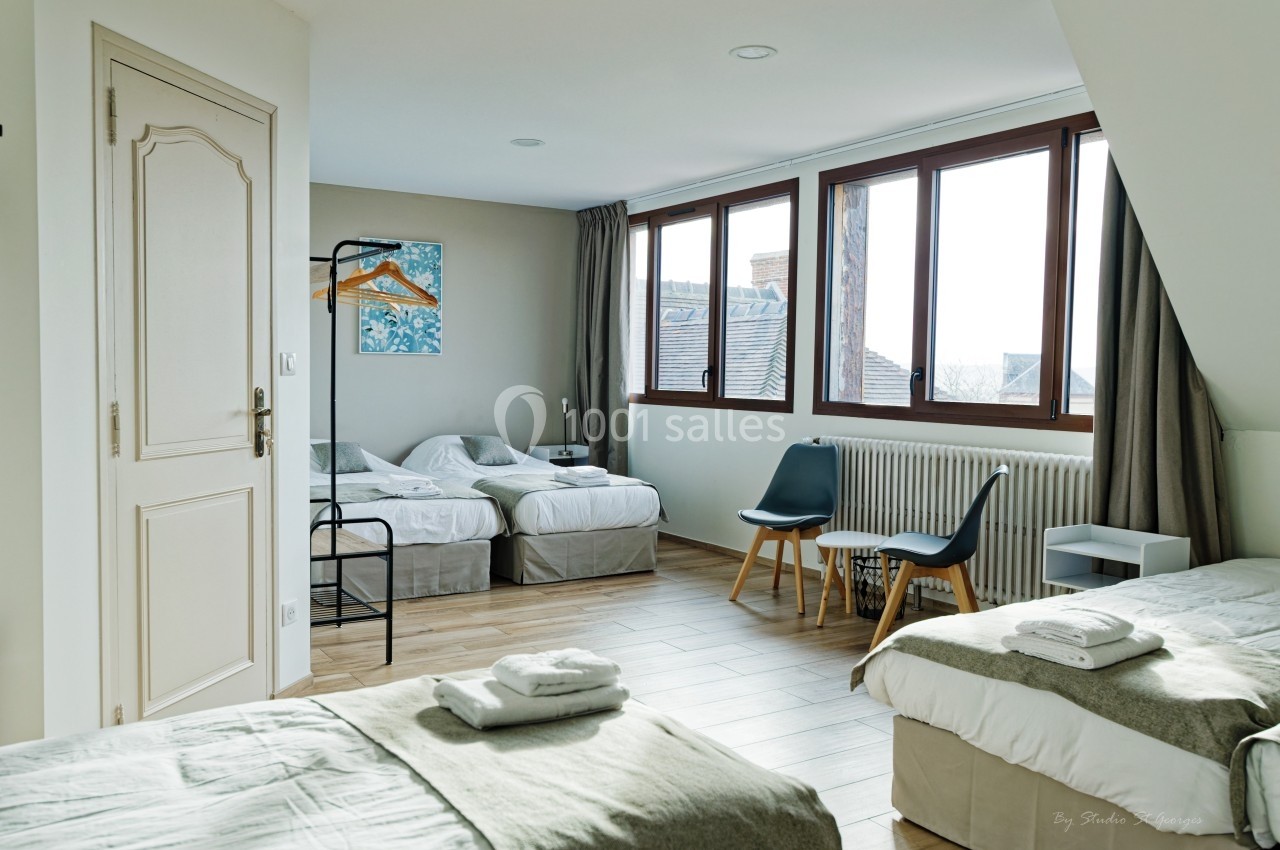 Chambre lumineuse avec plusieurs lits simples, grande fenêtre, chaises et porte-manteau dans un décor épuré.