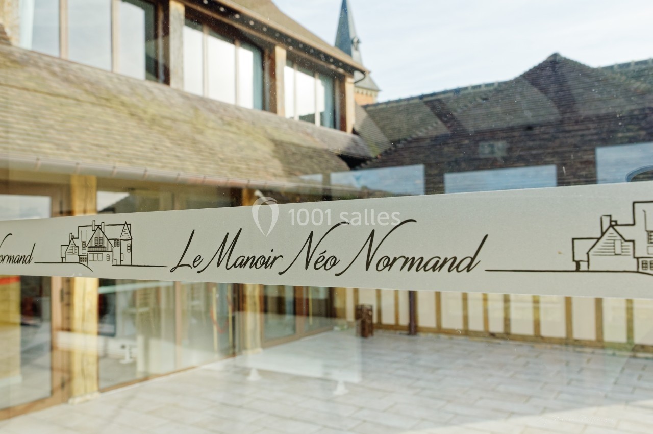 Vue d'une entrée vitrée avec l'inscription ’Le Manoir Néo Normand’ et des bâtiments en arrière-plan.