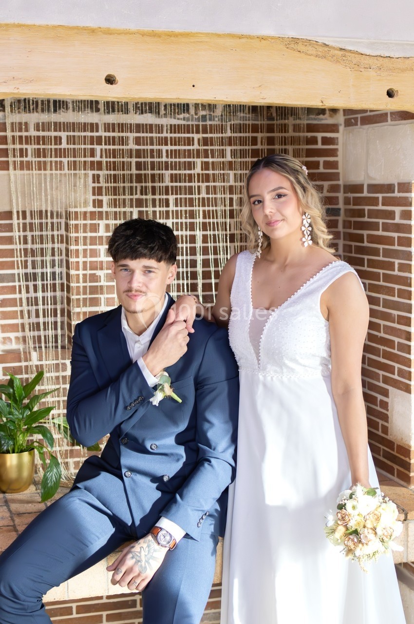 Un couple en tenue de mariage pose devant un mur en briques avec une plante verte à côté.