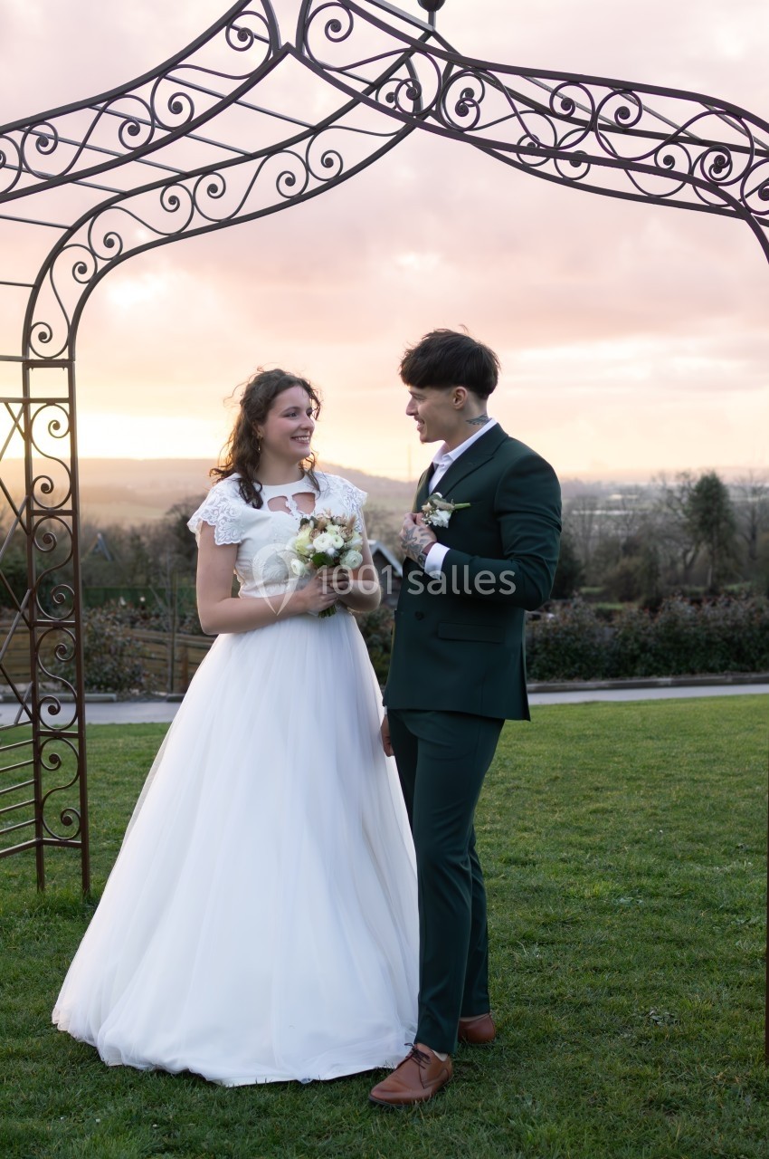 Un couple en tenue de mariage se tient sous une arche métallique dans un jardin au coucher du soleil.