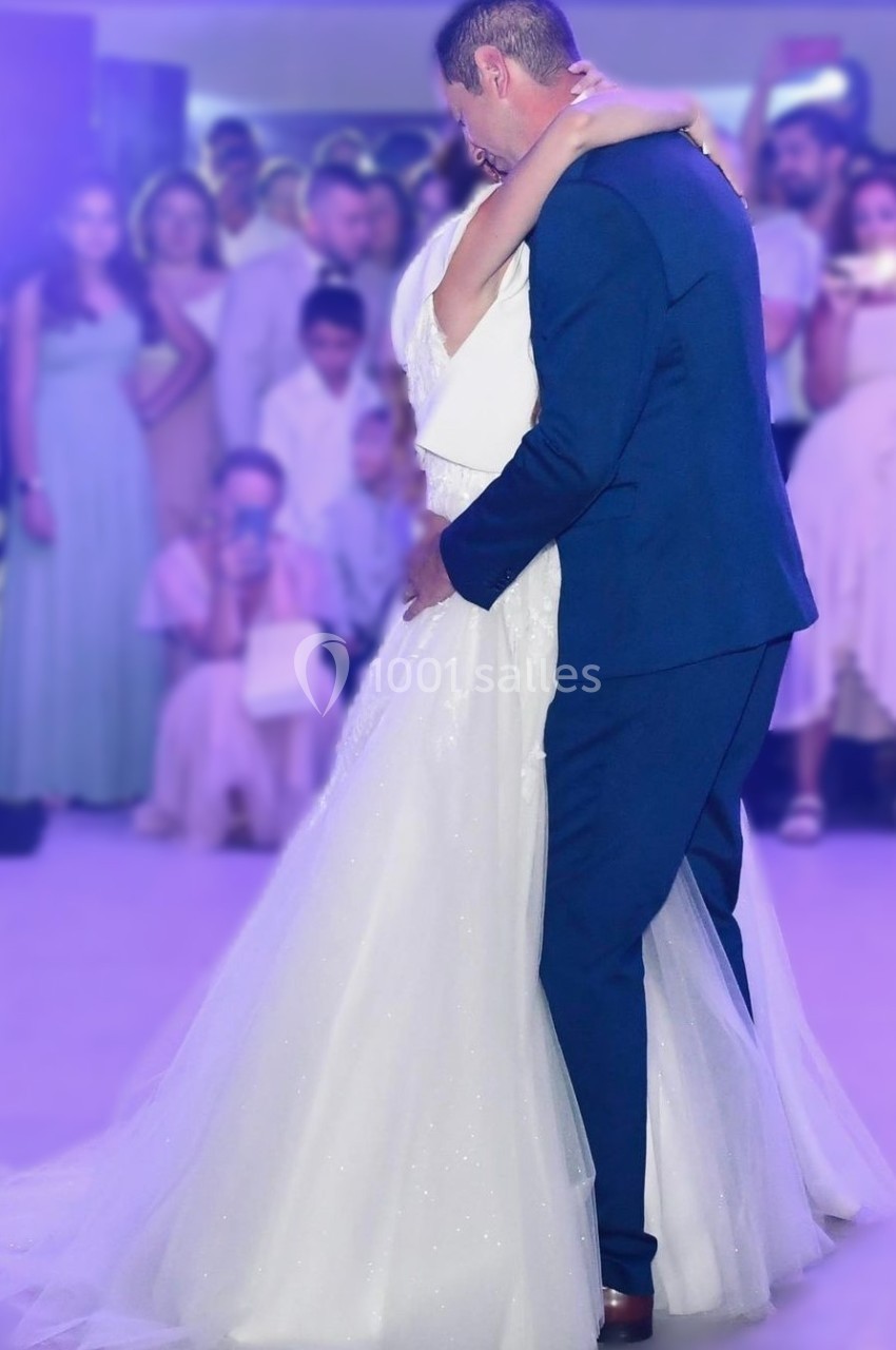 Un couple en tenue de mariage danse sous des lumières violettes, entouré d'invités en arrière-plan flou.