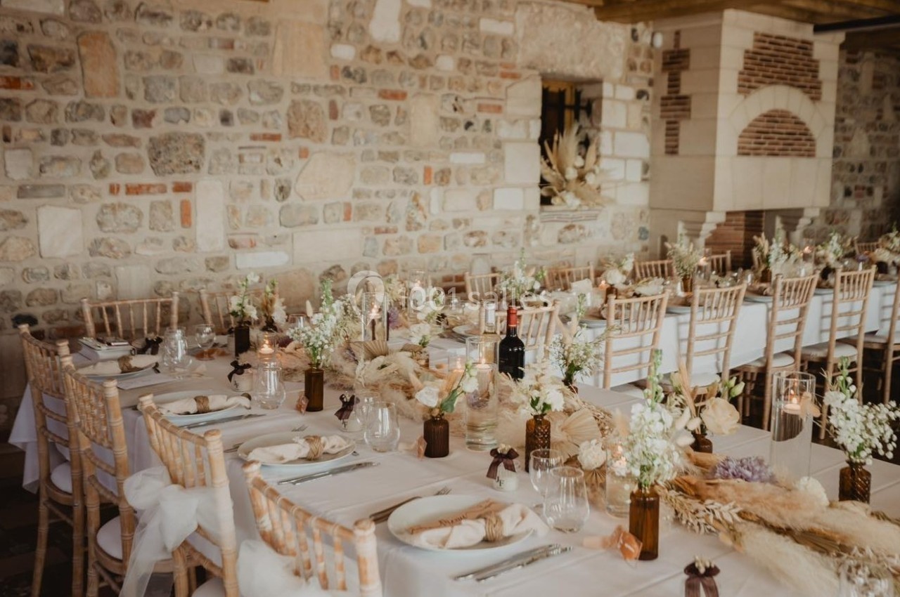 Salle décorée pour un repas festif avec tables dressées, chaises en bois, fleurs blanches et éléments naturels.