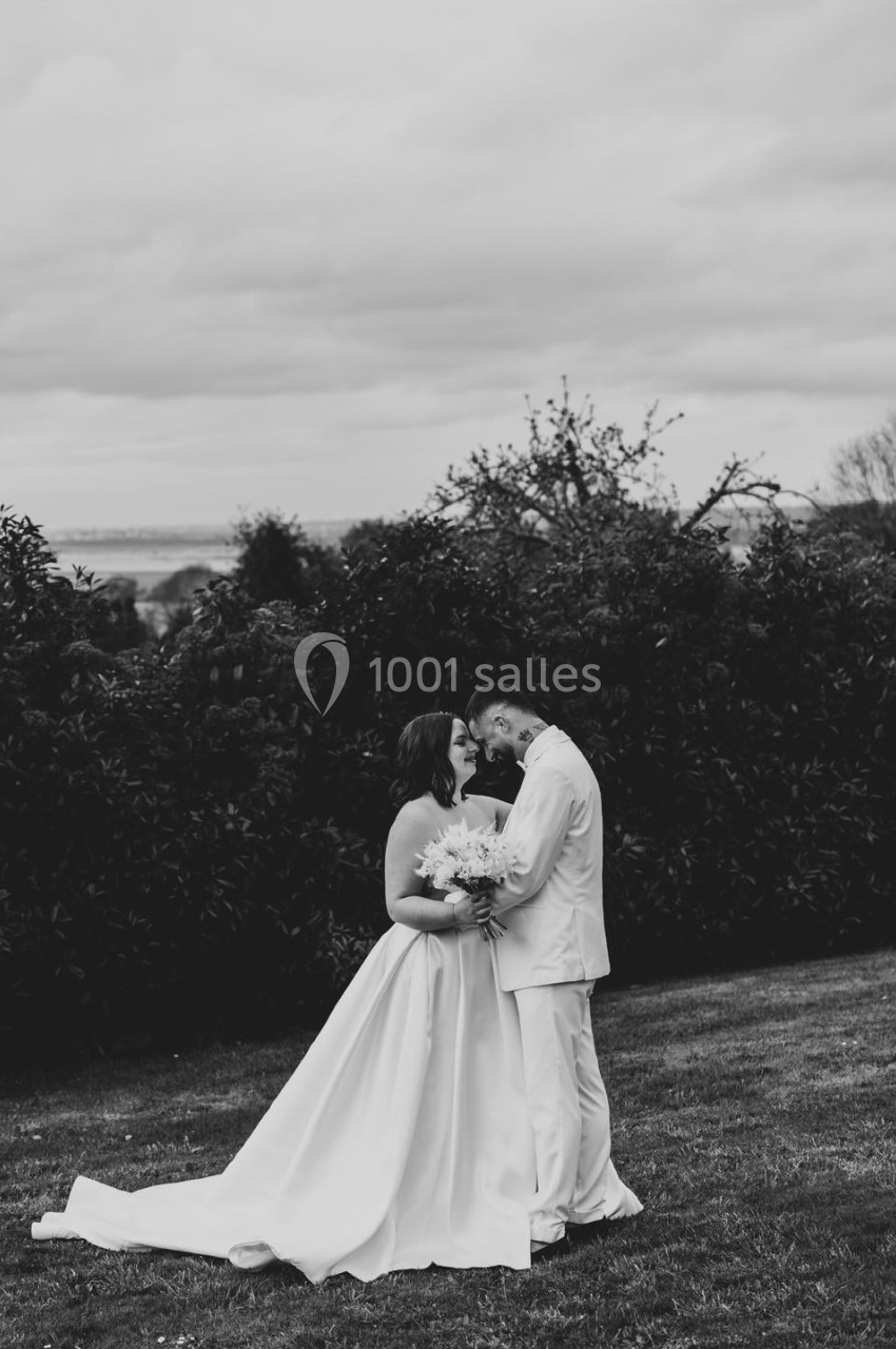 Un couple en tenue de mariage s'embrasse tendrement dans un jardin sous un ciel nuageux.
