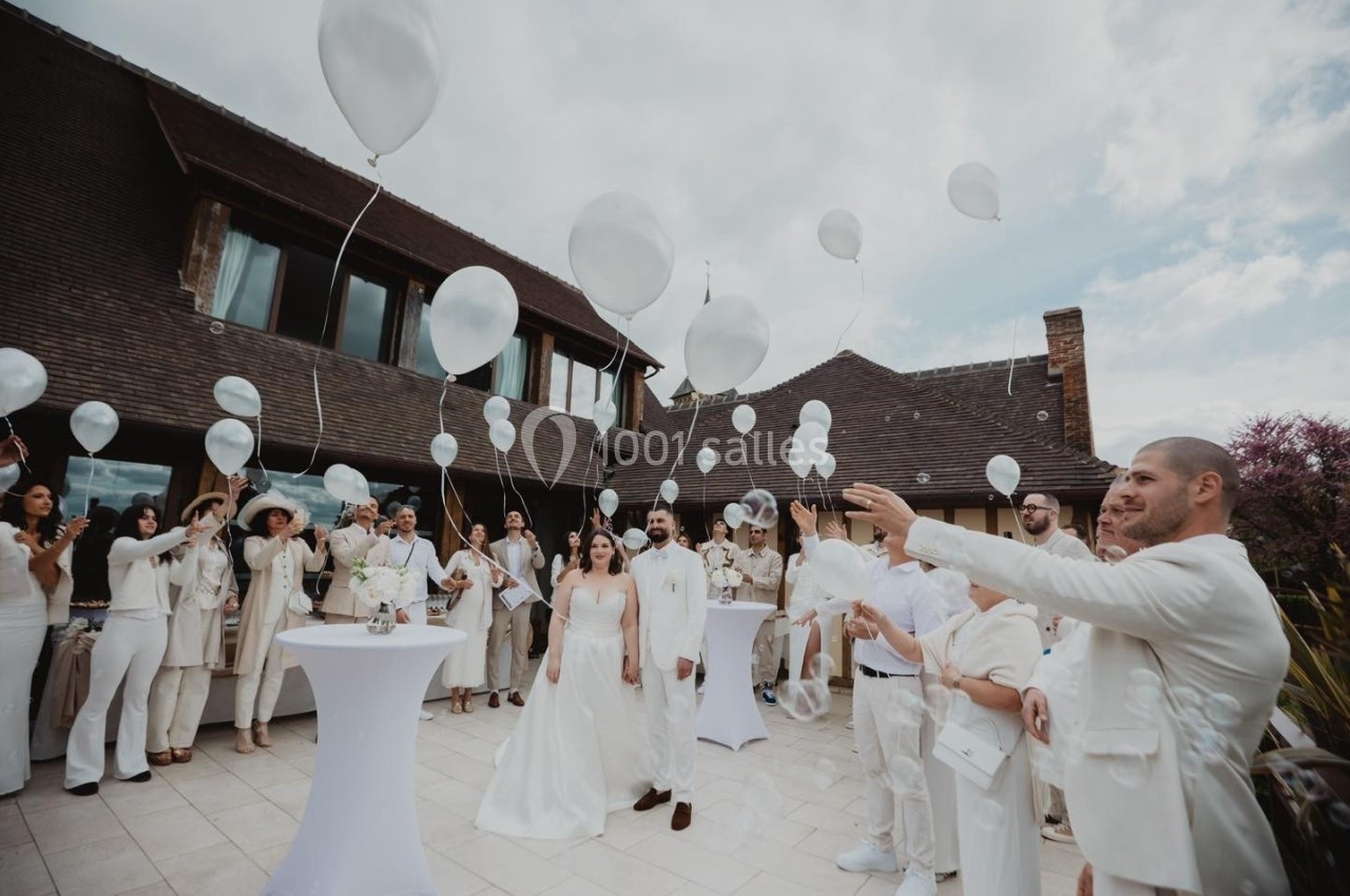 Des invités en tenue blanche lâchent des ballons blancs lors d'une célébration en extérieur devant un bâtiment en bois.