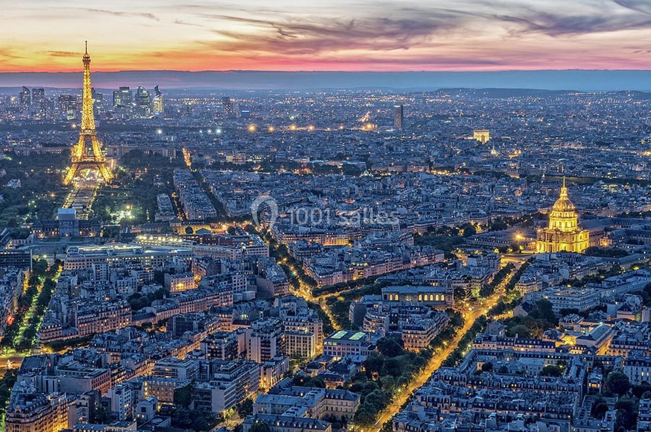 Vue aérienne de Paris au crépuscule, avec la tour Eiffel illuminée et les Invalides visibles au premier plan.