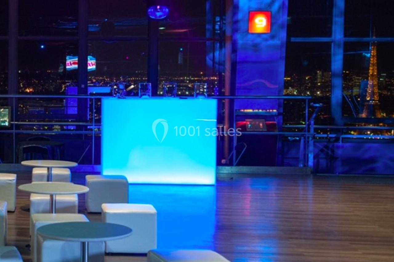 Vue intérieure d'un espace moderne avec un bar lumineux bleu, des sièges blancs et une vue nocturne sur la ville.