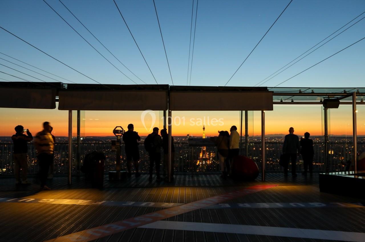 Personnes observant un coucher de soleil depuis une terrasse panoramique avec vue sur une ville illuminée.