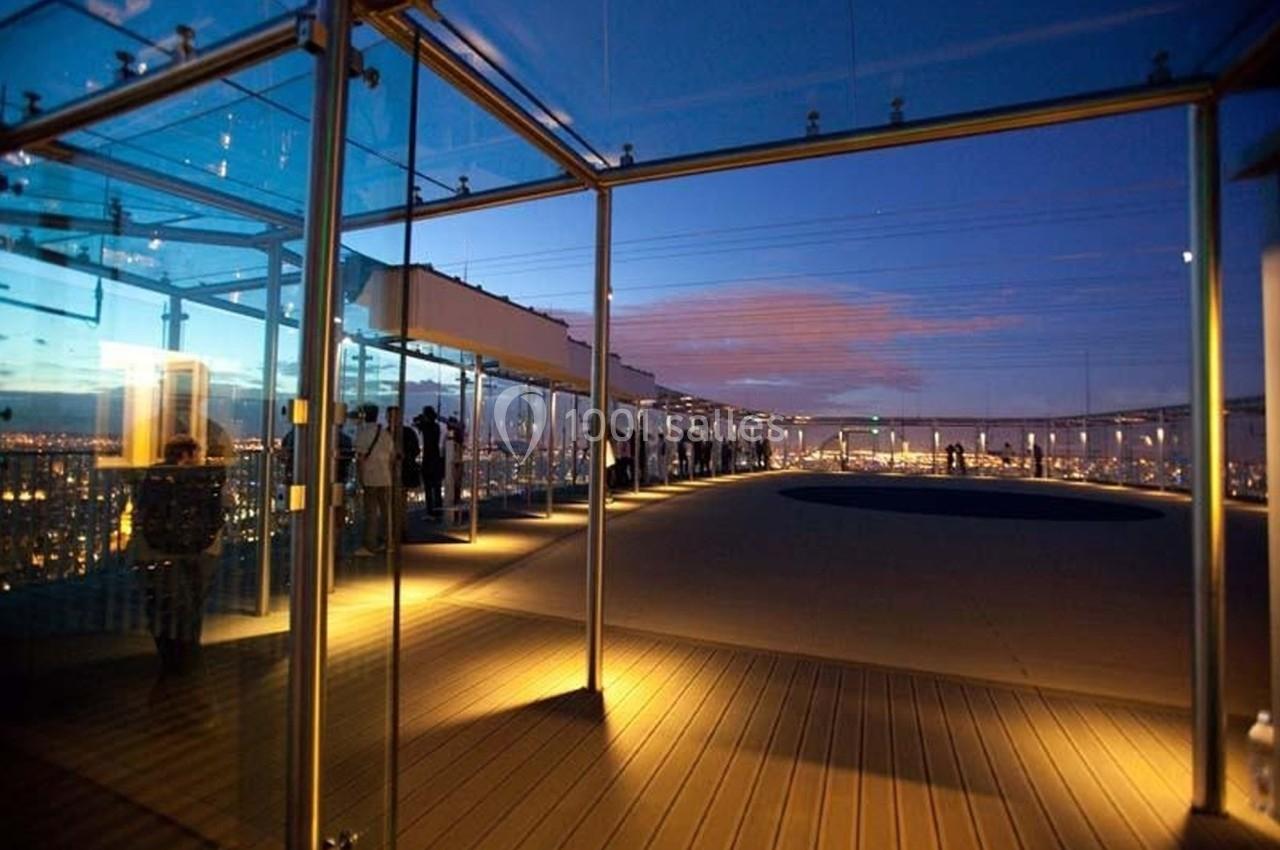Terrasse panoramique vitrée éclairée au crépuscule, avec des visiteurs observant la vue sur une ville illuminée.