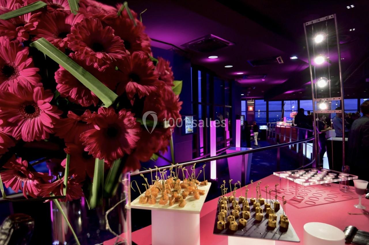 Bouquet de fleurs roses au premier plan avec un buffet de bouchées apéritives dans une salle éclairée en lumière violette.