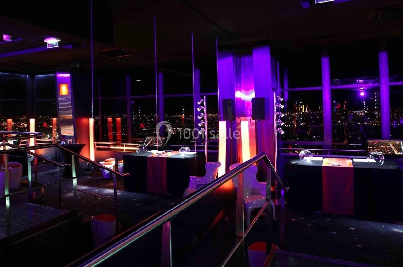 Salle sombre avec éclairage violet et rouge, tables décorées et vue nocturne sur une ville à travers de grandes fenêtres.