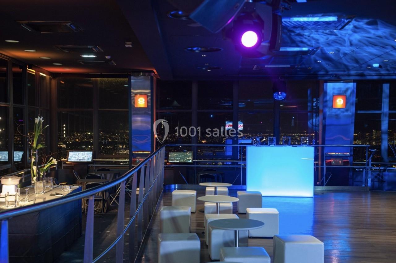 Salle moderne avec éclairage tamisé, tables basses blanches, bar et vue nocturne sur une ville illuminée.