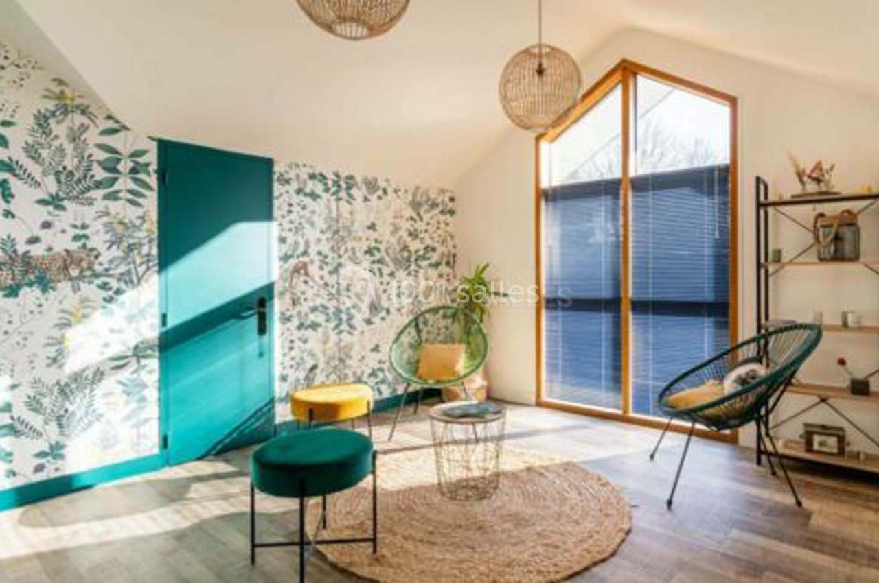 Salon lumineux avec mur végétal, grandes fenêtres, fauteuils modernes, tabourets colorés et étagère décorative.