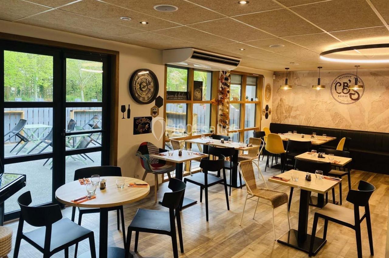 Salle de restaurant lumineuse avec tables dressées, chaises variées et vue sur une terrasse extérieure arborée.