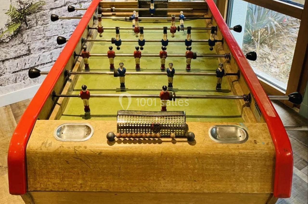 Baby-foot en bois avec joueurs rouges et bleus, situé dans une pièce lumineuse près d'une fenêtre et d'un mur en briques.