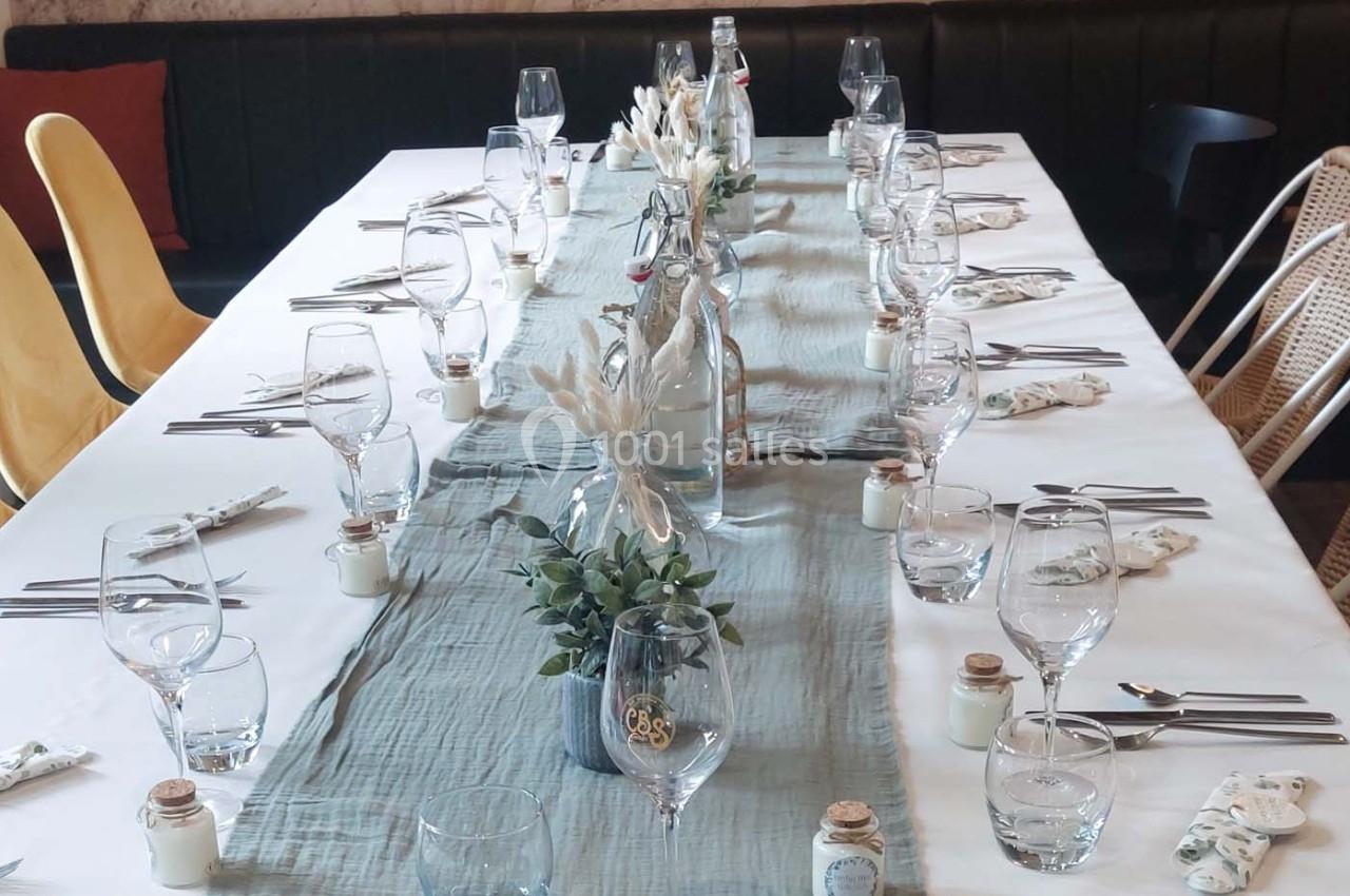 Table dressée avec une nappe blanche, chemin de table bleu, verres, couverts et décorations végétales.
