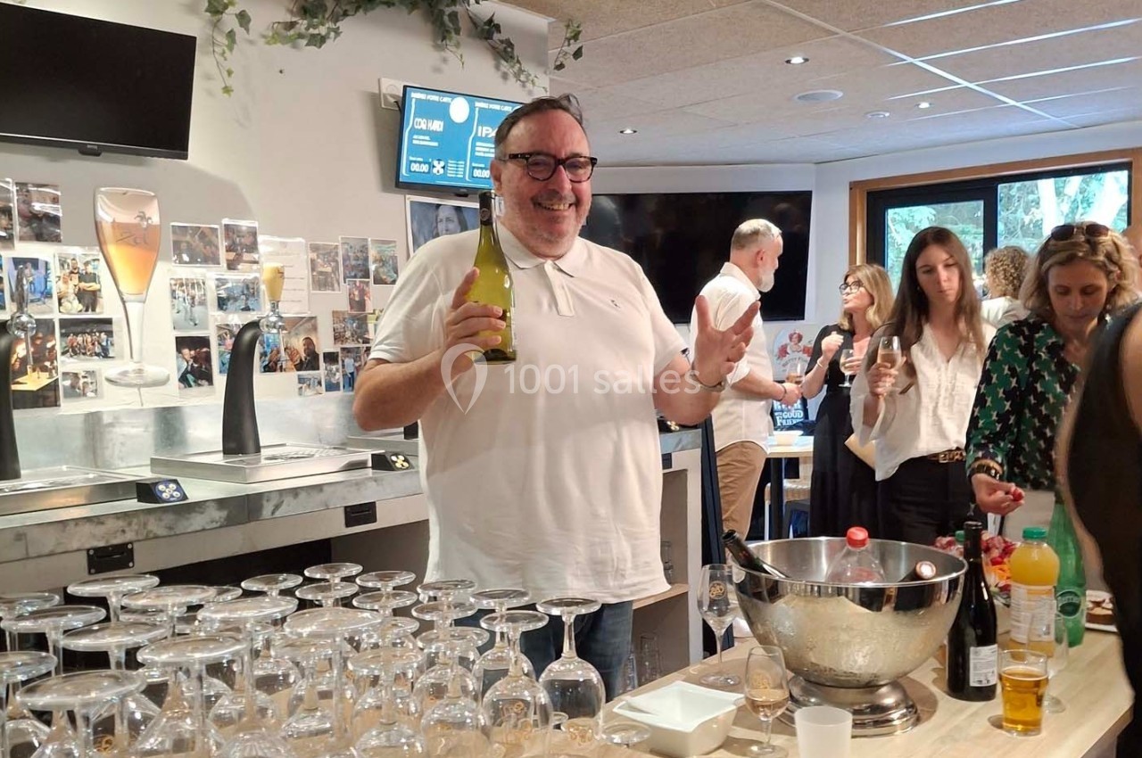 Un homme souriant tient une bouteille dans un bar lors d'un événement convivial, entouré de verres et de participants.