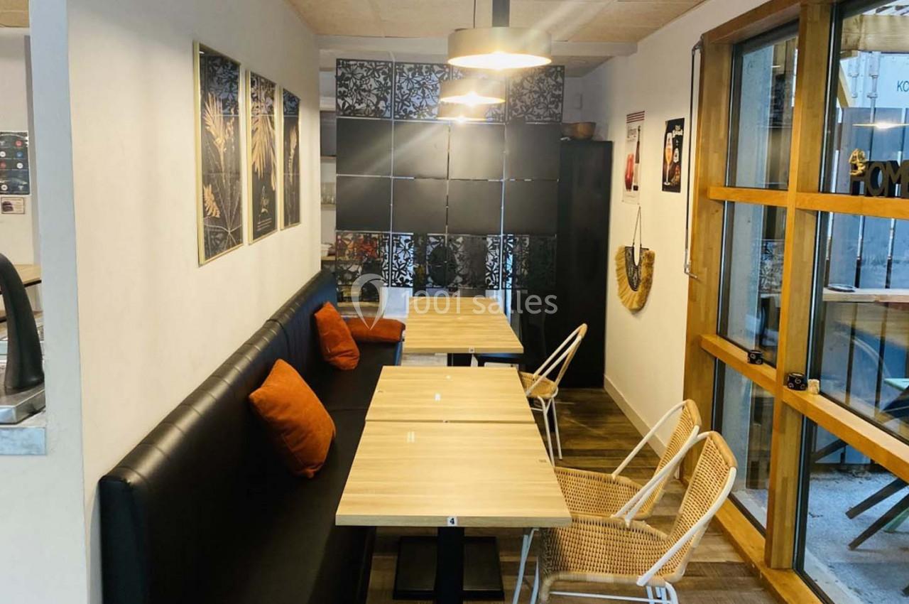 Salle de restaurant avec banquette noire, coussins orange, chaises en rotin et tables en bois clair près d'une baie vitrée.