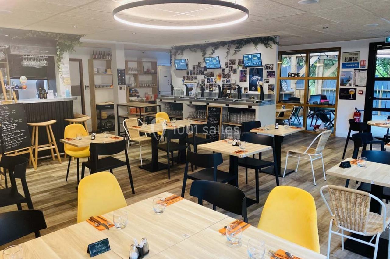 Salle de restaurant lumineuse avec tables en bois clair, chaises noires et jaunes, et un bar en arrière-plan.