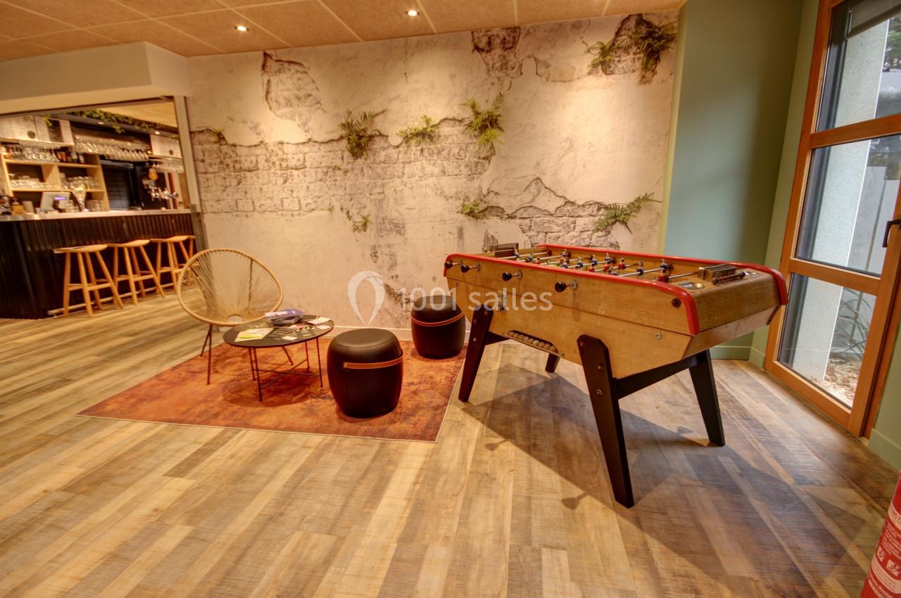 Espace détente avec baby-foot, fauteuil en rotin, poufs et mur décoratif végétalisé dans un intérieur chaleureux.