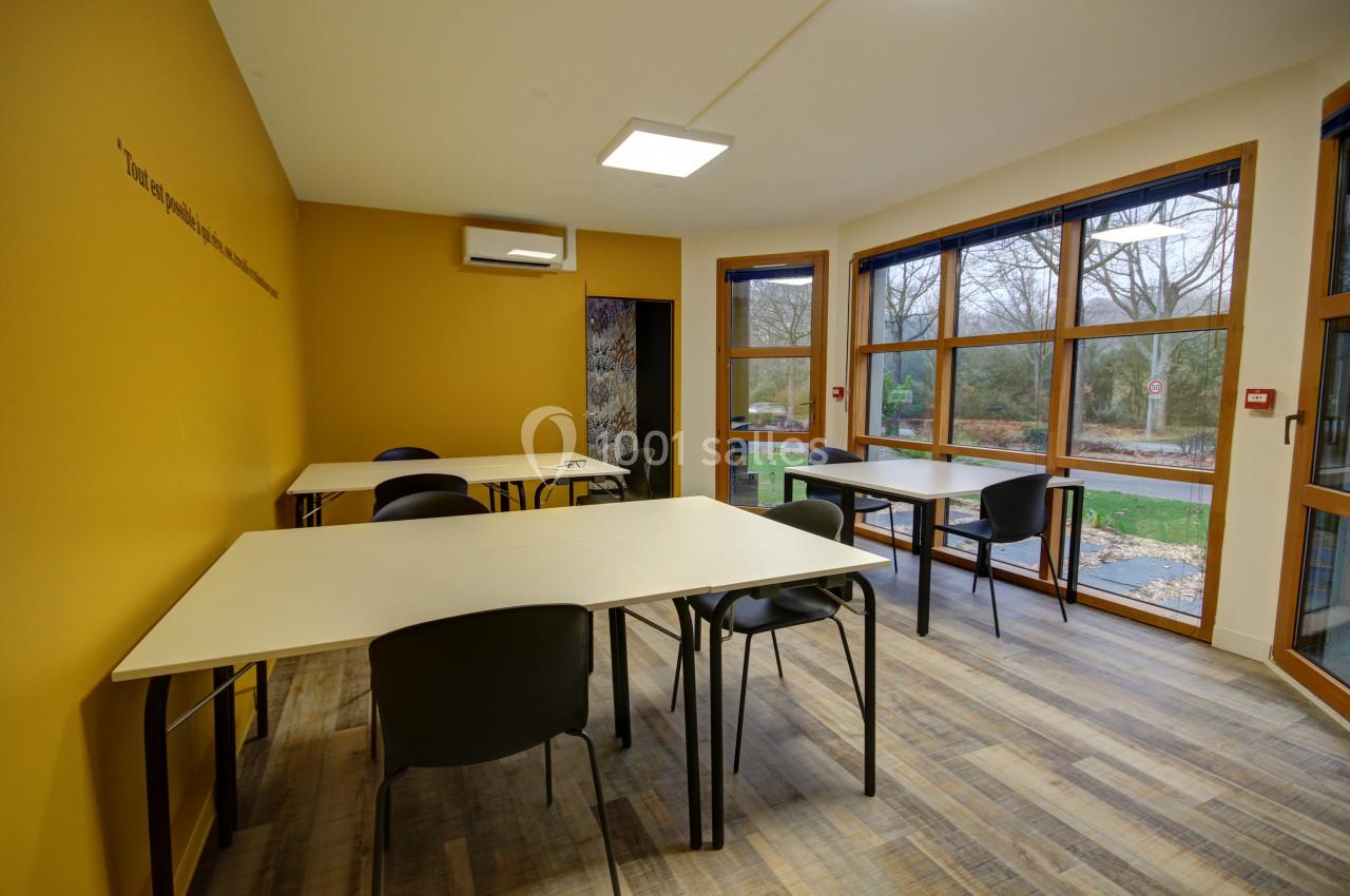 Salle lumineuse avec murs jaunes, tables et chaises noires, grandes fenêtres donnant sur un espace extérieur arboré.