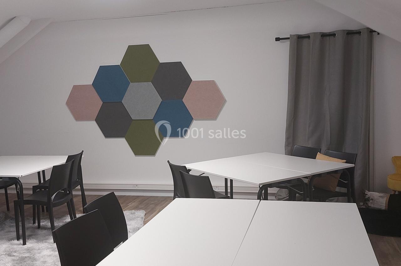 Salle lumineuse avec tables blanches, chaises noires, décoration murale hexagonale et coin détente avec fauteuil jaune.