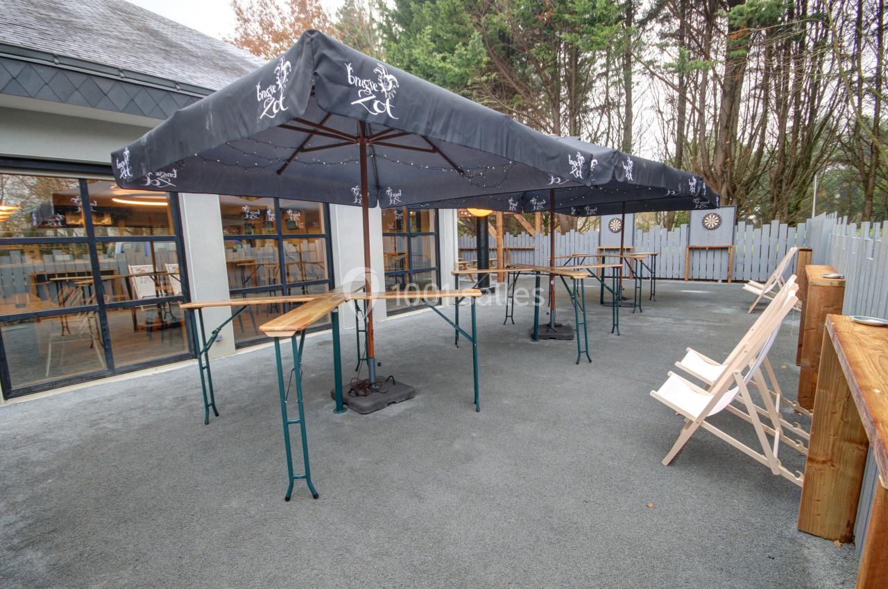 Terrasse extérieure avec tables hautes, parasols noirs et chaises longues en bois, entourée de végétation.