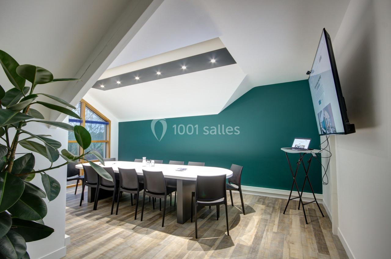 Salle de réunion moderne avec table ovale, chaises noires, mur vert, écran mural et plante en pot.