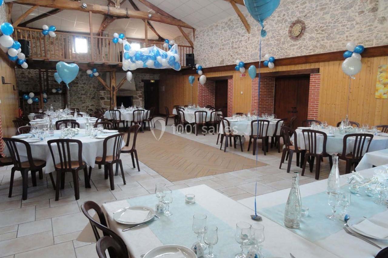 Salle de réception décorée avec des ballons bleus et blancs, tables dressées avec nappes claires et vaisselle.